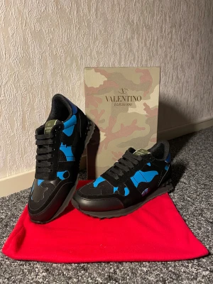 Valentino Garavani Rockrunner sneakers - Säljer ett par Valentino Garavani Rockrunner sneakers i svart och blått med coolt camouflage-mönster. Skorna har snörning, rund tå och detaljer i både mocka och skinn. Perfekta för dig som vill sticka ut med en lyxig streetwear vibe.