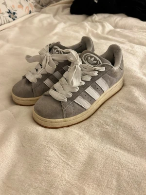 Grå Adidas Campus sneakers strl 37⅓ - Säljer ett par grå Adidas Campus sneakers med klassiska vita ränder och logga på tungan och hälen. Skorna har rund tå, platt sula och breda vita snören. Ovandelen är i mjuk mocka och insidan är fodrad i textil. Perfekt för dig som gillar retrostil och streetwear.