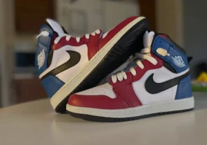 Nike Air Jordan 1 röd/blå/vit - Säljer ett par Nike Air Jordan 1 med högt skaft i en riktigt clean färgkombo av rött, blått, vitt och svart. Skorna har klassisk svart swoosh, vita snören och detaljerad Air Jordan-logga på sidan. Ovandelen är i skinn och sulan är svart och vit. Perfekta för dig som vill sticka ut.