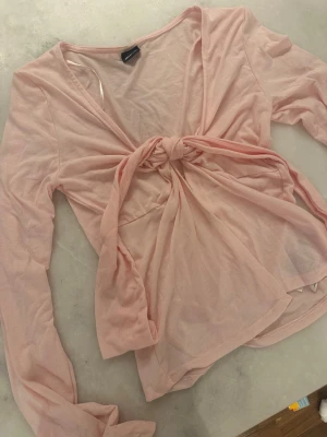 Ljusrosa omlottopp från Gina Tricot - Supersöt ljusrosa omlottopp från Gina Tricot med långa ärmar och knyt framtill. Tunt och lätt material som känns luftigt och perfekt för varmare dagar. Toppen har v-ringning och en feminin look som passar till jeans eller kjol.