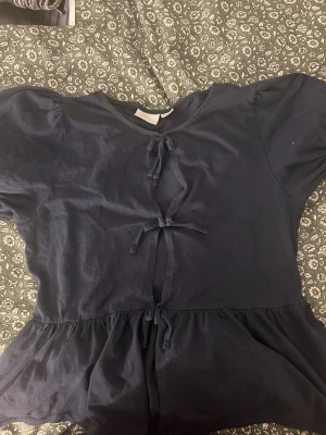 Mörkblå topp med knyt från Gina Tricot - Supersöt mörkblå topp från Gina Tricot med tre knyt framtill och volang nertill. Toppen har korta ärmar och är gjord i mjuk bomull, perfekt för en chill och trendig look.