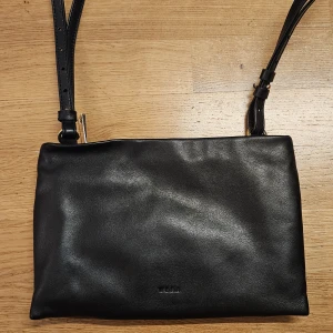 Svart axelväska i skinn från Wera - Nätt axelremsväska i läder med guldfärgade metalldetaljer. HELEN har en smal avtagbar och justerbar axelrem, och kan bäras som crossbody eller i handen som en kuvertväska. Väskan stängs med dragkedja och har ett stort fack, en ficka med dragkedja och en öppen ficka. Har använt enstaka gånger. Nypris 899 kr.   Mått: Längd: 25 cm Höjd: 17 cm Dropplängd från
