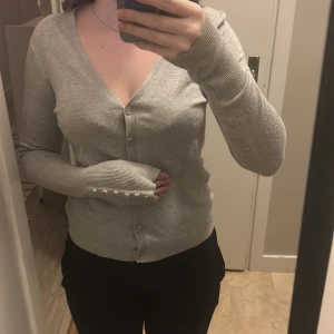 Ljusgrå kofta med knappar - Snygg ljusgrå kofta med v-ringning och långa ärmar. Koftan har ribbade muddar med dekorativa knappar längs ärmsluten och knäppning framtill. Perfekt för lager-på-lager och enkel att matcha med olika outfits. Den är lite nopprig