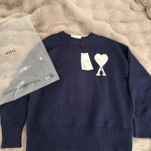 Mörkblå stickad tröja från Ami - Snygg mörkblå stickad tröja från Ami Alexandre Mattiussi med vit hjärtlogga på bröstet. Tröjan har rund hals, ribbade muddar och är tillverkad i mjuk bomull. Perfekt för dig som gillar stilrena och trendiga plagg med en twist.