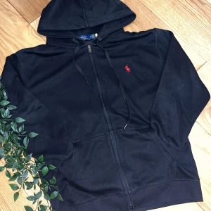 Svart hoodie från Polo Ralph Lauren - Svart hoodie från Polo Ralph Lauren med dragkedja och huva. Klassisk röd broderad logga på bröstet och känguruficka framtill. Tillverkad i mjuk bomull, perfekt för chill dagar eller när du vill ha en clean streetstyle.