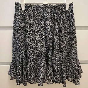 Svart leopardmönstrad kjol Retro Icone - Supersnygg kort kjol från Retro Icone med svart och vit leopardmönster. Kjolen har volangkant nedtill och ett elastiskt midjeband som gör den bekväm och enkel att bära.