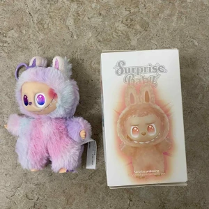 Surprise Baby Pretty Plush Pendant Blind Box - Söt och färgglad Surprise Baby plush pendant från blind box-serien. Dockan har mjuk päls i pastellfärger och stora glittrande ögon. Levereras med originalförpackning. Skicket är mycket bra, inga synliga defekter eller slitage. Perfekt som samlarfigur eller present.