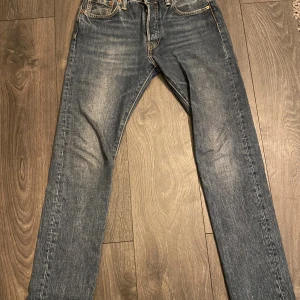 Levi's 501 jeans blå W29 L32 - Klassiska Levi's 501 jeans i blå tvätt med snygga slitningar. Modellen har rak passform, fem fickor och knappgylf. Jeansens material är robust denim med orange kontrastsömmar och ikonisk läderpatch bak. Perfekt för dig som gillar tidlös stil.