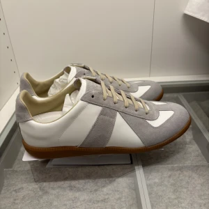 Maison Margiela GATs sneakers grå/vit - Maison Margiela GATs sneakers i vitt skinn och grå mocka med klassisk gummisula. Snygg retrostil med beige snörning och diskreta detaljer. Perfekta för dig som gillar minimalistisk och stilren design.