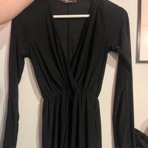 Svart omlottklänning från Gina Tricot - Svart långärmad klänning från Gina Tricot med omlott-skärning och markerad midja. Klänningen har djup v-ringning och är tillverkad i ett mjukt, stretchigt material som ger en snygg passform. Perfekt för dig som gillar stilrena och enkla plagg.