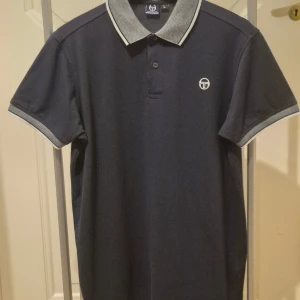 Marinblå vintage pikétröja Sergio Tacchini L - Snygg marinblå vintage pikétröja från Sergio Tacchini med kontrasterande grå och vit krage samt ärmslut. Klassisk design med två knappar och diskret logga på bröstet. Perfekt för dig som gillar sportig och stilren look.