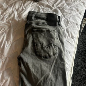 Replay Anbass grå jeans, storlek 32 - Snygga Replay Anbass jeans i grå tvätt med klassisk femficksdesign och svarta detaljer. Jeansen har rak passform och normal midja, tillverkade i mjukt jeanstyg med Replay-logga på bakfickan. Perfekta för dig som gillar stilrena och moderna jeans.