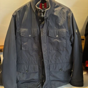 Mörkblå jacka från J.Lindeberg - Snygg mörkblå vindjacka från J.Lindeberg med hög krage, fyra framfickor med lock och knappar, samt dragkedja och knappstängning. Jackan har ett rött innerfoder och en patch på ärmen. Jackan är perfekt för hösten/Vintern, då den har relativt mycket innerfoder 