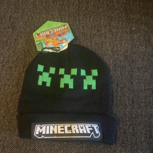 Svart Minecraft mössa med creeper-motiv - Svart mössa med gröna creeper-figurer och stor Minecraft-logga framtill. Perfekt för dig som älskar gaming och vill visa din stil även när det är kallt ute. Tillverkad i mjukt stickat material som håller dig varm.
