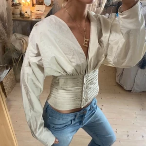 Beige blus med v-ringning - Säljer en beige korsettopp med bred v-ringning och långa ärmar. Toppen har en markerad midja med knappar framtill och ett plisserat tyg som ger en cool siluett. Skulle även passa M