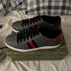 Gucci sneakers med GG-mönster - Snygga Gucci sneakers i grått med klassiskt GG-mönster, svarta skosnören och vit sula. Sidan har röd och marinblå rand, och baksidan har metallicröd och metallicblå detalj med Gucci-logga. Perfekta för dig som vill sticka ut med lyxig streetstyle.