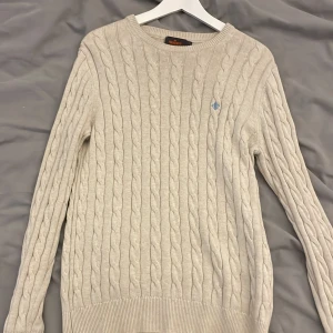 Beige kabelstickad tröja Morris S - Snygg beige kabelstickad tröja från Morris i storlek S, tillverkad av 100% pima bomull. Tröjan har ett diskret blått broderat emblem på bröstet, rund hals och ribbade muddar vid ärmslut och nederkant. Perfekt för dig som gillar klassisk stil. Och den blir perfekt för vintern. Hör bara av dig om du har frågor🤗