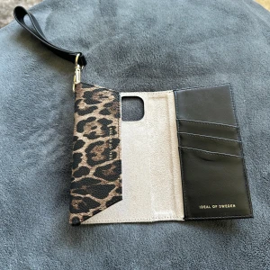 Leopardmönstrad plånbok IPhone 13 från IDEAL OF SWEDEN - Snygg plånbok till IPhone 13 med leopardmönster och svarta detaljer från IDEAL OF SWEDEN. Plånboken har flera kortfack, magnetstängning och en praktisk handledsrem i svart. Materialet är skinnimitation med lyxig känsla och guldiga detaljer på remmen