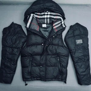 Svart dunväst från Burberry med huva - Svart dunväst från Burberry med avtagbara ärmar och stor huva. Insidan av huvan har klassiskt rutigt Burberry-mönster i grått, vitt och rött. Västen har dragkedja, knappar och två fickor med dragkedja framtill. Märkespatch på ena ärmen och insidan.