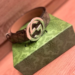 Gucci GG bälte med silverdetalj - Säljer ett Gucci GG bälte i brunt skinn med klassiskt GG-mönster och stor silverfärgad logga som spänne. Bältet har beige insida och kommer med originalbox, dustbag, kvitto och kontrollkort. Perfekt accessoar för att lyfta din outfit.