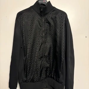 Svart bomberjacka från BOSS - Snygg svart bomberjacka från BOSS med diskret rutmönster på framsidan och baksidan. Jackan har hel dragkedja, ribbad krage och muddar. Materialet är en mix av polyester och bomull som ger en skön känsla och stilren look.