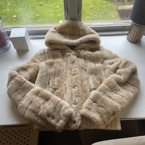 Beige pälsjacka från Juicy Couture - Säljer en beige pälsjacka från Juicy Couture i storlek S. Jackan har huva, guldfärgade detaljer och knappar framtill. Insidan är quiltad med broderad logga och kedja vid nacken. Perfekt för dig som vill ha en lyxig och cozy vibe i höst och vinter. Nypris 5000