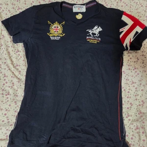 Svart Geographical Norway t-shirt - Cool svart t-shirt från Geographical Norway med broderade detaljer och Union Jack-flagga på ena ärmen. Snygga tryck och broderier både fram och bak, samt röda sömmar längs sidorna. V-ringad hals och korta ärmar. Perfekt för dig som gillar sportig och unik stil.