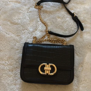 Svart axelväska  - Snygg svart axelväska med mönster. Väskan har både kedja och justerbar axelrem i skinn, samt ett praktiskt innerfack med dragkedja. Perfekt storlek för det viktigaste och ger en lyxig vibe till din outfit. Väskan är precis som ny!