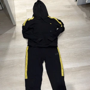 Svart hoodie/byxa från EA7 med gula detaljer Full set - Svart hoodie/byxa från EA7 Emporio Armani med gula ränder längs axlarna/benen där det står EA7. Tröjan har långa ärmar och ribbade muddar. Perfekt för en sportig och avslappnad look