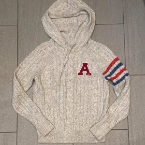 Stickad hoodie från Abercrombie & Fitch - Grå stickad hoodie från Abercrombie & Fitch med kabelstickat mönster, huva med snörning och röd A-logga på bröstet. Ena ärmen har ränder i rött, blått och vitt. Perfekt för höst och vinter när du vill vara både varm och snygg.