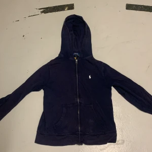 Mörkblå hoodie från Polo Ralph Lauren - Snygg mörkblå hoodie från Polo Ralph Lauren med dragkedja och huva. Tröjan har ett klassiskt vitt Polo-logo broderat på bröstet, två fickor fram och är tillverkad i mjuk bomull. Perfekt för chill dagar och enkel att matcha med jeans eller joggers.