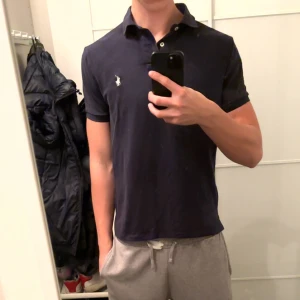 Mörkblå piké från Polo Ralph Lauren - Säljer en mörkblå pikétröja från Polo Ralph Lauren med vit broderad logga på bröstet. Modellen är Custom Slim Fit med klassisk krage och knappar framtill. Perfekt för dig som gillar stilren och tidlös design.
