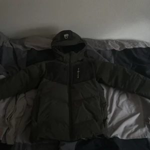 Grön Sail racing Antarctica pufferjacka - Säljer en mörkgrön pufferjacka från Sail racing med luva och svart panel upptill. Jackan har broderad logga på bröstet och en badge på ärmen. Fylld med syntetmaterial och det saknas en liten del av dragkedjan men det är inget problem. Perfekt för kalla dagar.