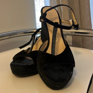 Svarta kilklack sandaler med T-rem - Snygga svarta sandaler från Unisa med kilklack och öppen tå. Skorna har en T-rem med glittriga detaljer och justerbar ankelrem. Ovandelen är i mocka och sulan har en flätad look. Perfekta för dig som gillar höga klackar och stilren design.