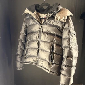 Moncler grå pufferjacka med huva - Snygg grå pufferjacka från Moncler med glansig finish, huva och två stora dragkedjefickor framtill. Jackan har en klassisk Moncler-logga på ärmen och ribbad krage. Perfekt för kalla dagar när du vill hålla stilen.Går även att ta av luvan ifall man vill.Hör gärna av er om ni undrar något