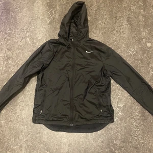 Svart Nike vindjacka med huva - Svart vindjacka från Nike med huva och dragkedja framtill. Jackan har två fickor med dragkedja, diskret Nike-logga på bröstet och meshfoder på insidan. Perfekt för löpning eller utomhusaktiviteter. Lätt och smidig design med justerbara ärmslut. Passar nån som är 155-165cm