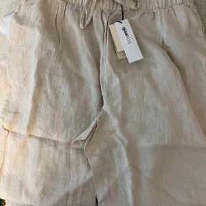 Beiga linnebyxor från Gina Tricot - Snygga beiga shorts från Gina Tricot i 100% linne. De har elastisk midja med snörning för skön passform och en avslappnad look. Perfekta för varma dagar och ger en clean vibe till din outfit.
