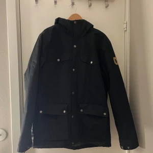 Svart vinterjacka  från Fjällräven  - Svart parkajacka från Fjällräven med huva och fyra fickor framtill. Jackan har knappar och dragkedja, samt ett mysigt foder på insidan. Perfekt för kalla dagar och outdoor vibes. Klassisk och stilren design som funkar till det mesta. Nypris 3999 kr! Använt fåtal gånger, inga defekter. Skriv för fler bilder