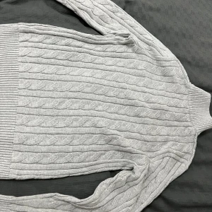 Ljusgrå stickad polotröja med half zip - Säljer en ljusgrå stickad polotröja med kabelstickat mönster och ribbade muddar. Tröjan har hög krage med half zip-dragkedja och långa ärmar. Perfekt för kyliga dagar och riktigt snygg till jeans eller kjol.