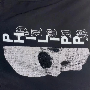 Svart hoodie med döskalle - Svart hoodie med dragkedja, huva och fickor. Framsidan har en stor glittrig döskalle och coolt tryck med logga. Märkets emblem i gummi på bröstet. Perfekt för dig som gillar streetstyle och vill sticka ut.