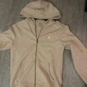 Beige hoodie från Polo Ralph Lauren - Säljer en beige hoodie från Polo Ralph Lauren med dragkedja och klassisk huva. Tröjan har fickor framtill och den ikoniska vita loggan broderad på bröstet. Perfekt för chill dagar och snygg till jeans eller joggers. Använd fåtal gånger