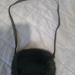 Svart fluffig axelväska med dragkedja - Säljer en rund, svart axelväska med fluffig pälsimitation och guldfärgad dragkedja. Väskan har ett långt axelband i syntetmaterial och är perfekt för att bära dina viktigaste grejer. Enkel och cool design som sticker ut.