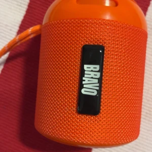 Bravo Bluetooth-högtalare - Portabel Bravo Bluetooth-högtalare i snygg orange färg. Kompakt design med bärrem, perfekt för musik på språng. Ytan ser ut att vara i mycket gott skick utan synligt slitage eller repor. Lätt att ta med och enkel att använda.