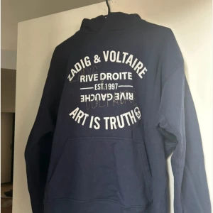 Mörkblå hoodie Zadig & Voltaire - Mörkblå hoodie från Zadig & Voltaire med vit text och coola detaljer i strass framtill. Klassisk känguruficka, huva och lång ärm. Snyggt tryck med 'ART IS TRUTH' och döskalle, perfekt för en edgy streetstil.