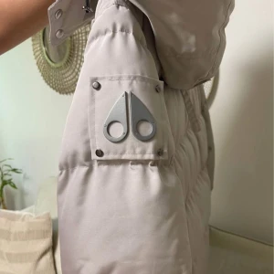 Beige dunjacka från Moose Knuckles - Säljer en beige dunjacka från Moose Knuckles med ikonisk logga på ärmen, svarta detaljer och dragkedjor. Jackan har huva med svart fuskpäls, flera fickor med knapp och zip, samt snygga slitsar med dragkedja i sidorna. Perfekt för kalla dagar och riktigt stilren.