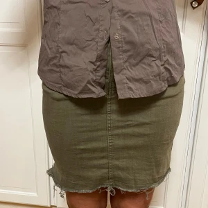 Olivgrön jeanskjol med rå kant - Snygg olivgrön jeanskjol med rå, fransig kant nedtill och klassiska jeansdetaljer som knapp och fickor. Kjolen har en rak, tight passform och är sydd i ett slitstarkt bomullsmaterial. Perfekt för dig som gillar en enkel men cool stil.
