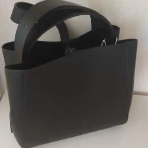 Svart axelväska i skinn med dragkedja - Stilren svart axelväska i skinn med breda handtag och en praktisk dragkedja upptill. Väskan har en minimalistisk design med slät yta och rymligt fack, perfekt för att bära med sig allt man behöver. Snygg och enkel att matcha med olika outfits.