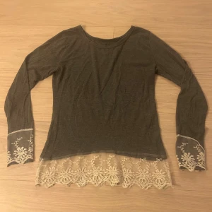 Grå långärmad topp med spetsdetaljer - Supersnygg grå långärmad topp med ljus spets och broderade detaljer vid ärmslut och nederkant. Toppens spets ger en romantisk och unik vibe, perfekt för dig som gillar att sticka ut lite extra. Mjuk och skön passform med rund halsringning.