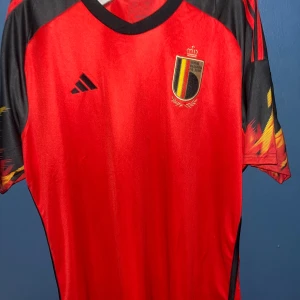 Belgien matchtröja Adidas 2XL - Säljer en officiell Belgien landslagströja från Adidas i storlek 2XL. Tröjan är röd med svarta och gula detaljer på ärmarna, har korta ärmar och broderat landslagsemblem på bröstet. Materialet är lätt polyester, perfekt för fotboll eller häng med polarna. Skriv gärna till mig om ni har några frågor. Har aldrig haft på mig den