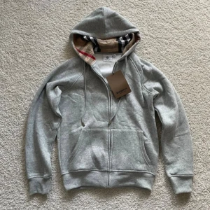 Grå hoodie från Burberry med dragkedja - Snygg grå hoodie från Burberry med klassiskt rutigt foder i huvan och dragkedja framtill. Tröjan har snörning i huvan, känguruficka och ribbade muddar. Perfekt för dig som vill ha en stilren och lyxig vardagslook. Hör gärna av er vid frågor 😊
